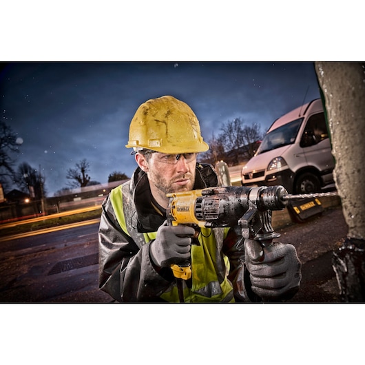 A person wearing a yellow hard hat and reflective safety vest uses a DEWALT electric drill outdoors, with a van and blurred lights visible in the background. The person's face is obscured.