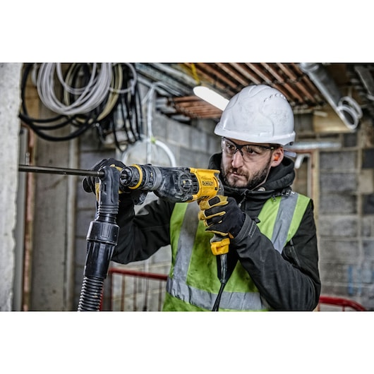 A person wearing a safety helmet and reflective vest is using a DEWALT D25134K rotary hammer drill in a construction setting with concrete and electrical cables visible.