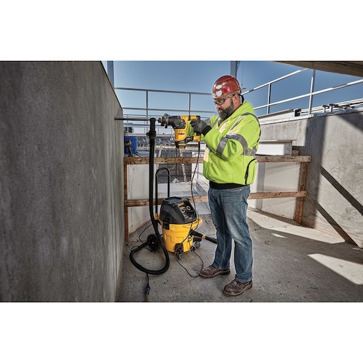 A person wearing a high-visibility jacket and a red hard hat is using a DEWALT power tool to drill into a concrete wall at a construction site. A yellow and black DEWALT vacuum is connected to the tool to manage dust.