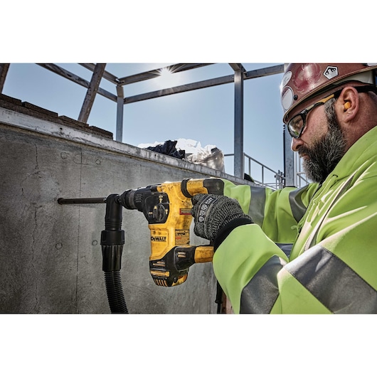 A person wearing a high-visibility safety jacket and gloves is using a yellow DEWALT rotary hammer drill to bore a hole into a concrete wall at a construction site. The person's face is blurred for privacy.