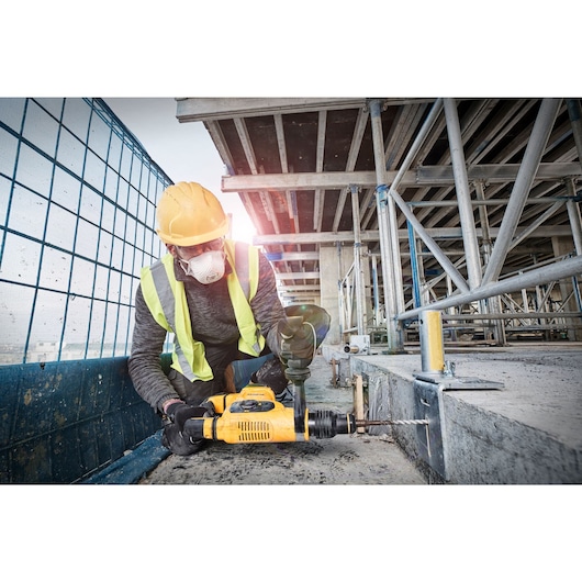 A construction worker wearing a yellow hard hat and high-visibility vest is using a yellow DEWALT rotary hammer drill (model D25810K) to drill into concrete at a building site.