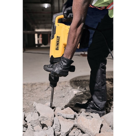 A person wearing black gloves and work boots is using a yellow DEWALT demolition hammer to break concrete rubble at a construction site.
