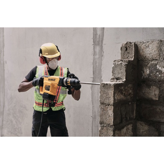 A construction worker wearing safety gear, including a helmet, ear protection, gloves, and a mask, uses a yellow DEWALT demolition hammer to break through a concrete wall.