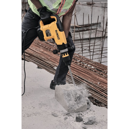 A worker using a DEWALT electric demolition hammer to break a concrete block at a construction site. The worker is wearing gloves and safety clothing, with rebar and unfinished structure visible in the background.