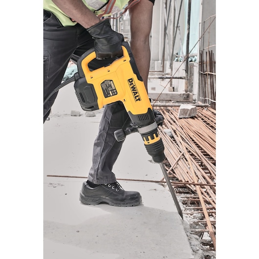 A person wearing protective gloves and boots uses a yellow DEWALT power tool to break concrete at a construction site. Steel rebar and building materials are visible in the background.