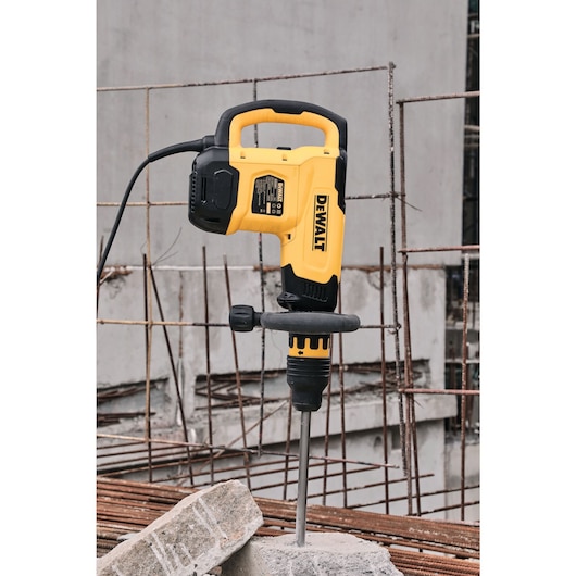 A DEWALT electric demolition hammer is positioned upright with its chisel point inserted into a concrete block at a construction site. Metal rebar and unfinished concrete structures are visible in the background.