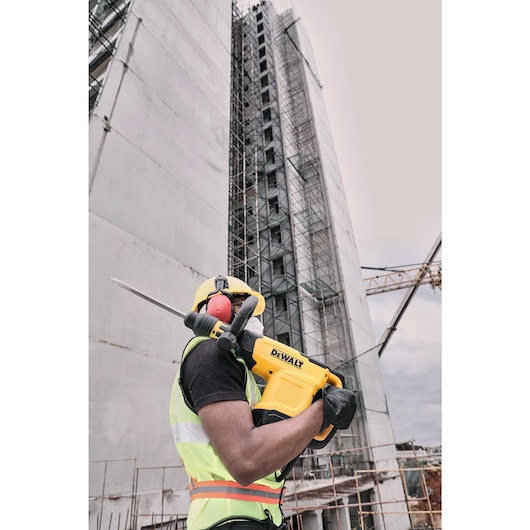 A construction worker wearing safety gear holds a yellow DEWALT demolition hammer (model D25881K) at a building site, with tall scaffolding and a crane in the background.