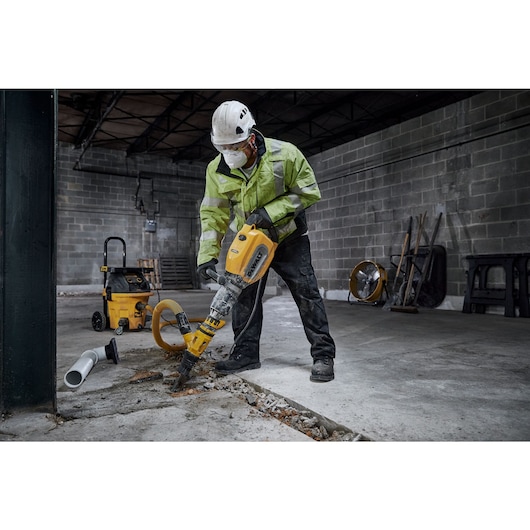 A construction worker wearing safety gear uses a yellow DEWALT D25911 demolition hammer to break concrete in an industrial setting.