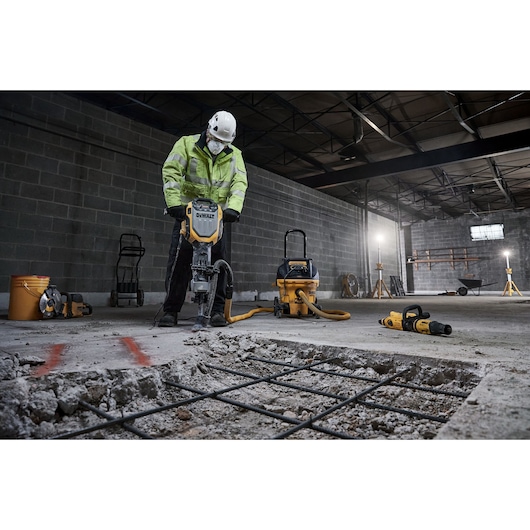 A worker wearing a safety helmet and a high-visibility jacket uses a DEWALT demolition hammer to break up concrete in an industrial setting. Various construction tools and equipment, including a vacuum and power tools, are visible in the background.
