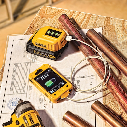 A DEWALT portable battery charger (DCB090) is shown charging a smartphone with a USB cable on a work table that has copper pipes and engineering plans. The battery charger displays charging progress, and the smartphone is in a yellow rugged case.