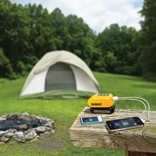 A DEWALT portable battery charger is placed on a wooden surface outdoors, with two smartphones connected and charging. In the background, there is a tent set up on grass and a stone firepit, with a forest and blue sky visible.