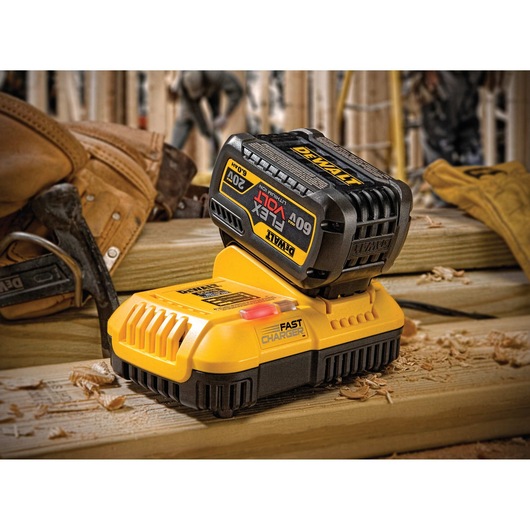 A DEWALT fast charger with a lithium-ion battery pack is placed on a wooden work surface. The charger and battery appear in a woodworking setting, with gloves and tools visible in the background.