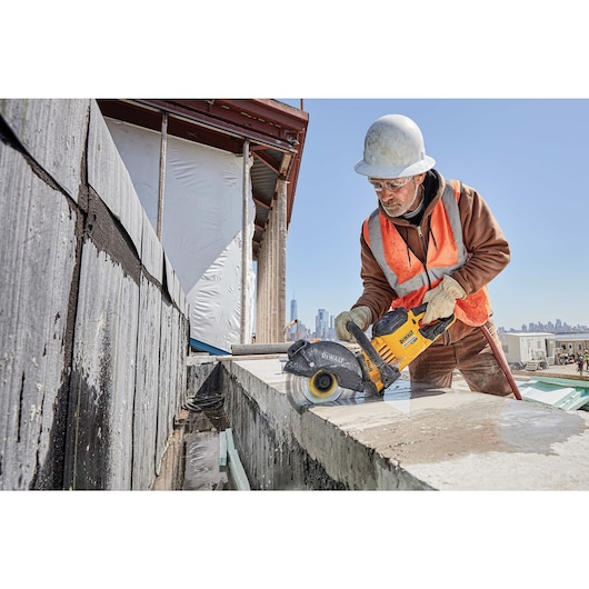 A construction worker wearing a hard hat and safety vest is using a yellow DEWALT DCB612 power tool to cut through a concrete block at an outdoor building site. The worker's face is blurred for privacy.