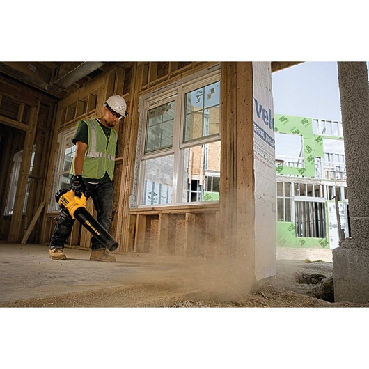 A construction worker wearing a safety helmet and reflective vest uses a yellow DEWALT cordless leaf blower to clear dust inside a building under construction.