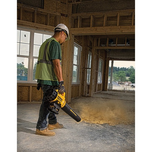 A person wearing safety gear uses a DEWALT cordless blower to clear dust from a concrete floor in a construction site.