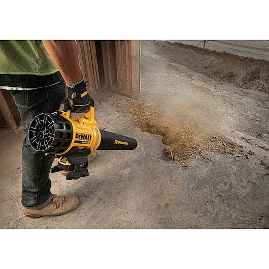 A person operating a yellow DEWALT cordless leaf blower to clear dust and debris from a concrete floor at a construction site.