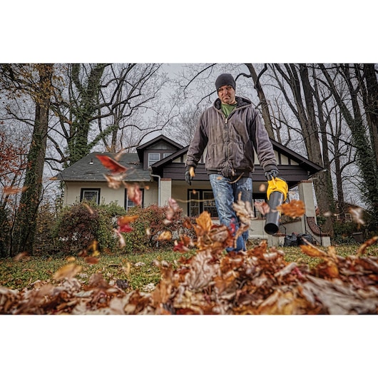 A person using a DEWALT leaf blower in front of a house, clearing leaves from the lawn. The person is wearing a jacket, jeans, gloves, and a hat. The focus is on the leaf blower and the flying leaves.