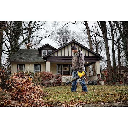 A person is using a yellow DEWALT leaf blower to move leaves in the yard outside a house, surrounded by trees in autumn.