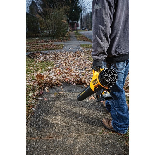 A person wearing a jacket and jeans is holding a DEWALT cordless leaf blower (model DCBL720B) while standing on a sidewalk covered with fallen leaves.