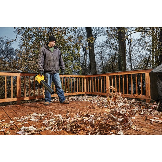 Person standing on a wooden deck using a yellow DEWALT leaf blower to clear fallen autumn leaves.