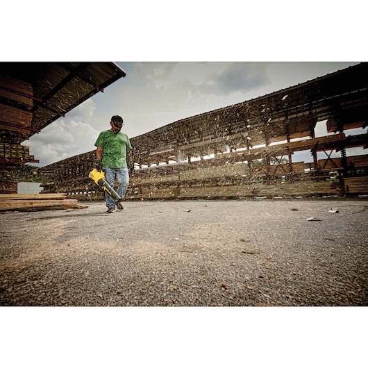 A person wearing a green shirt and jeans is using a yellow DEWALT leaf blower to clear debris on an outdoor surface in an industrial area with stacks of wooden planks.