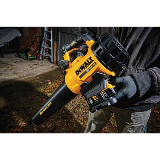 A person wearing gloves is holding a yellow and black DEWALT 20V cordless leaf blower, model DCBL720B, outdoors near a shed with gardening tools visible in the background.