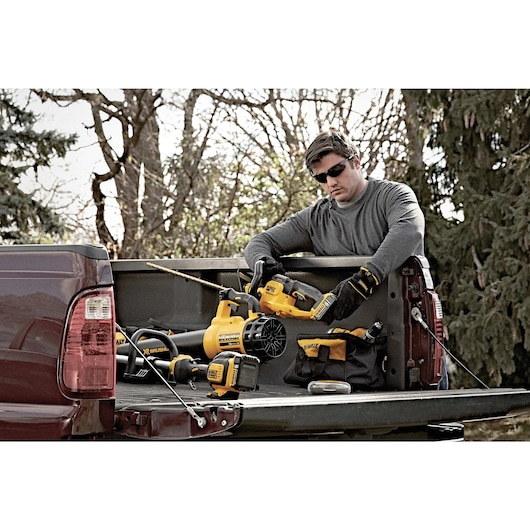 A person with a blurred face, wearing gloves and a long-sleeve shirt, is standing at the open tailgate of a pickup truck outdoors. On the truck bed are several yellow and black DEWALT power tools, including a leaf blower, a drill, and a tool bag.