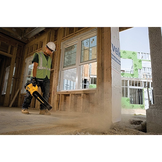 A construction worker wearing a safety helmet and reflective vest uses a yellow DEWALT cordless leaf blower to clear dust inside a building under construction.