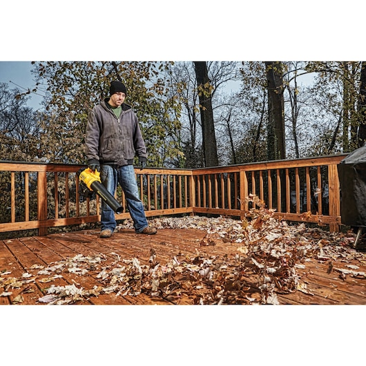 Person standing on a wooden deck using a yellow DEWALT leaf blower to clear fallen autumn leaves.