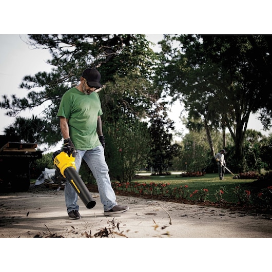 A person wearing gloves and outdoor clothing is using a DEWALT cordless leaf blower to clear debris from a walkway in a garden setting. Another person is working in the background among trees and plants.