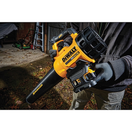 A person wearing gloves is holding a yellow and black DEWALT 20V cordless leaf blower, model DCBL720B, outdoors near a shed with gardening tools visible in the background.