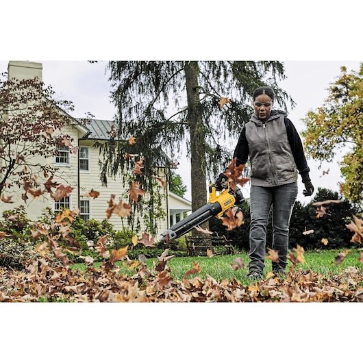 A person using a DEWALT cordless leaf blower (model DCBL722P1) in a yard, blowing leaves across the grass in front of a house.