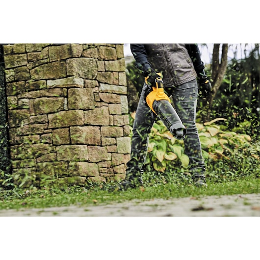 A person using a DEWALT leaf blower outdoors, blowing grass and leaves near a stone wall and garden plants.