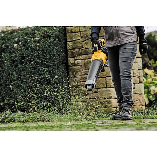 A person uses a yellow DEWALT cordless leaf blower outdoors to clear leaves and debris from the ground, with a stone wall and shrubbery in the background.