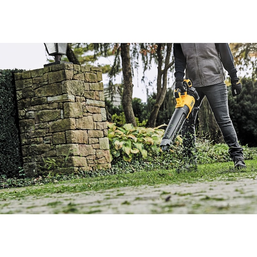 A person using a DEWALT cordless leaf blower outside on a garden path, blowing leaves and debris. The person is wearing gloves and a jacket, and part of a stone wall is visible in the background.