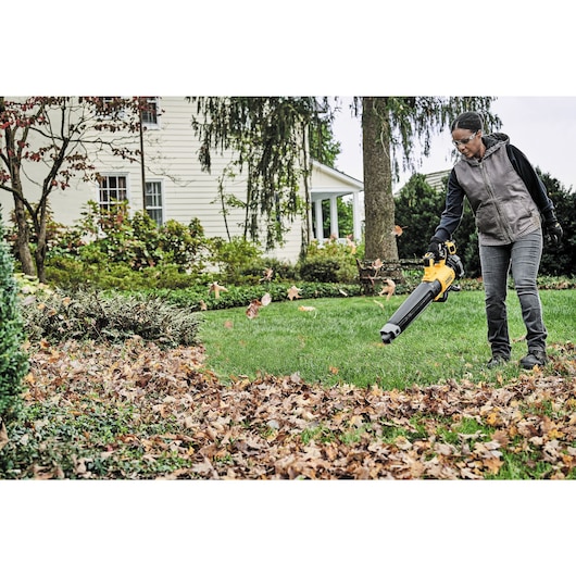 A person uses a DEWALT DCBL722P1 cordless leaf blower to clear fallen leaves from a lawn in front of a house.