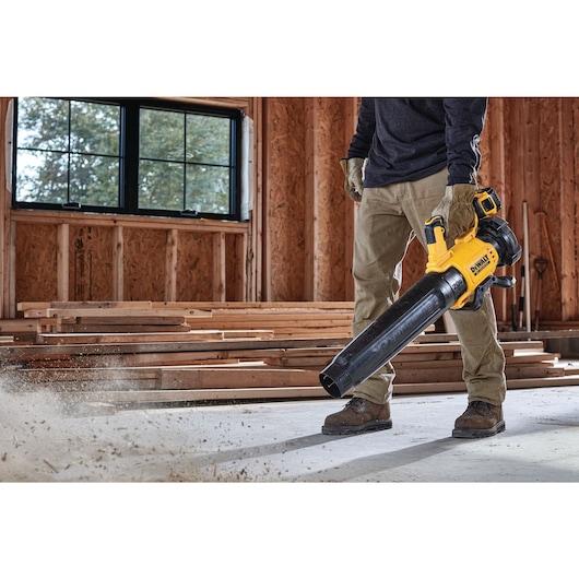 A person uses a DEWALT DCBL722P1 cordless blower to clear dust and debris from the floor of a construction site with unfinished wooden walls and stacked lumber.