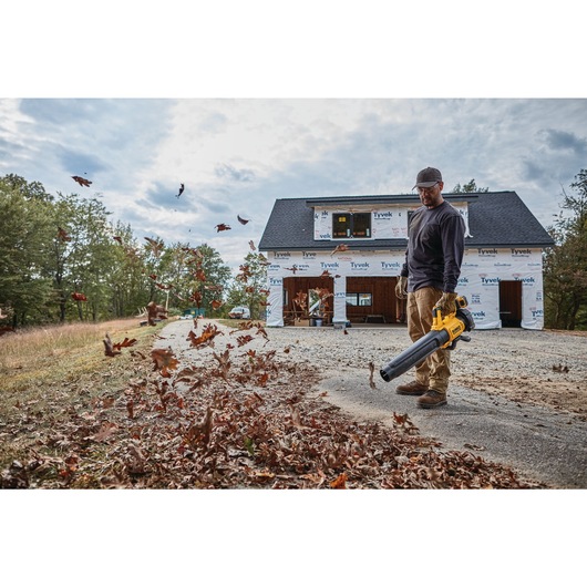 A person uses a yellow DEWALT leaf blower to clear leaves from a driveway in front of a house under construction.