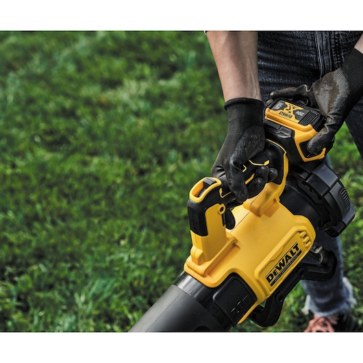 Close-up of a person outdoors using a DEWALT cordless leaf blower in a grassy area. The individual is wearing black gloves and holding the tool with both hands.