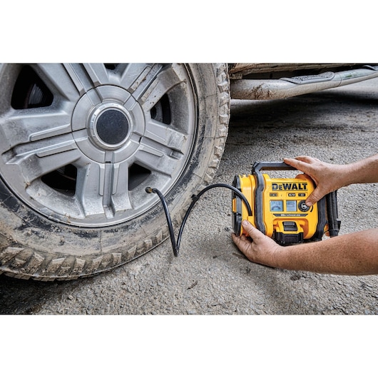 A person is using a DEWALT portable air inflator to inflate a dirty car tire on a paved surface.