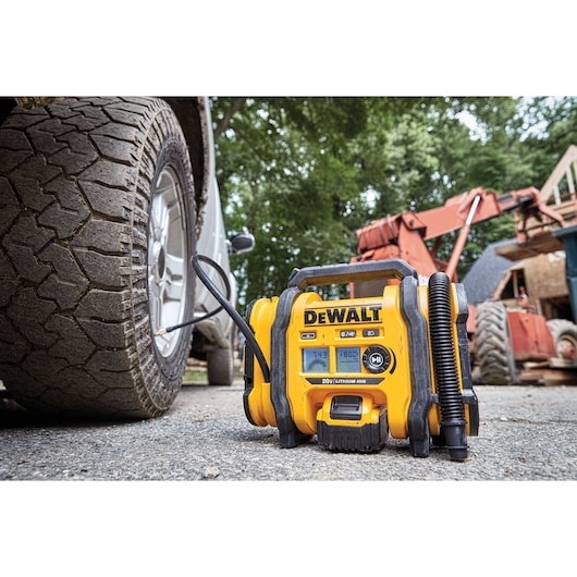 A DEWALT portable air inflator (model DCC020IB) is shown in use, inflating a vehicle tire outdoors. Construction equipment and a tree-filled background are visible.
