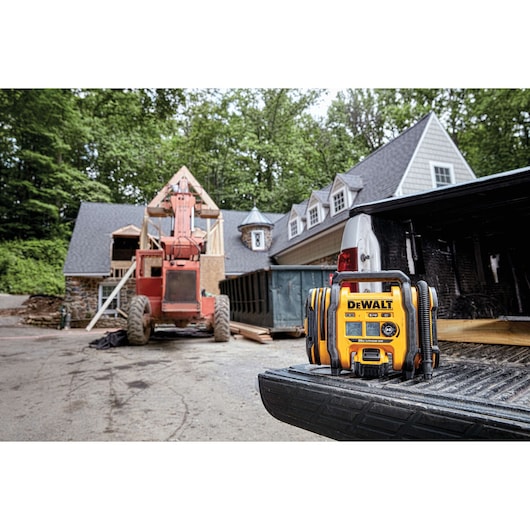 A DEWALT portable radio and charger is placed on the open tailgate of a pickup truck at a construction site, with a partially built house and construction equipment visible in the background.