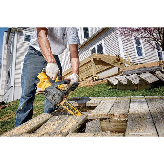 A person using a DEWALT cordless chainsaw (model DCCS620B) to cut wooden planks outdoors, with a house and deck construction in the background.