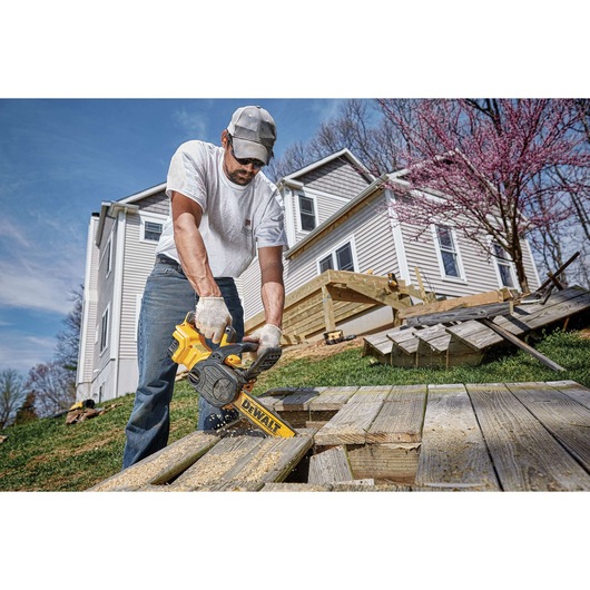 A person outdoors using a yellow DEWALT cordless chainsaw to cut wooden boards. The person is wearing gloves, a white shirt, jeans, and a cap. A light-colored house and blooming trees are visible in the background.