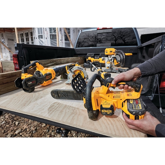 A person is holding a DEWALT cordless chainsaw on the bed of a pickup truck, surrounded by various other DEWALT power tools including a circular saw and a blower, all arranged on a sheet of plywood. The background shows a construction site with wooden beams and partially built structures.