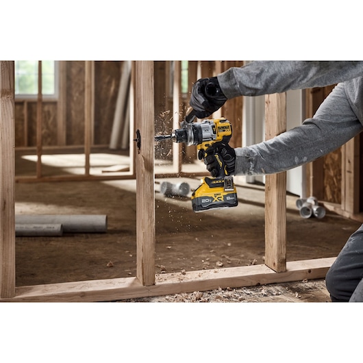 A person is using a DEWALT cordless drill to make a hole in a wooden stud inside a construction site. Wood shavings are visible as the drill operates, and the person is wearing gloves and a gray long-sleeve shirt.