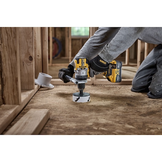 A person is using a DEWALT cordless power drill with a hole saw attachment to cut into a wooden subfloor inside a construction site with exposed wooden framing.