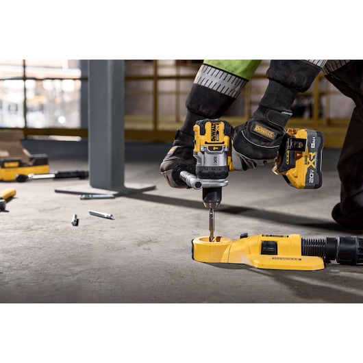 A person wearing gloves uses a DEWALT cordless drill to drill into a concrete floor, with a DEWALT dust extractor attachment connected. Various tools and bolts are scattered on the ground.