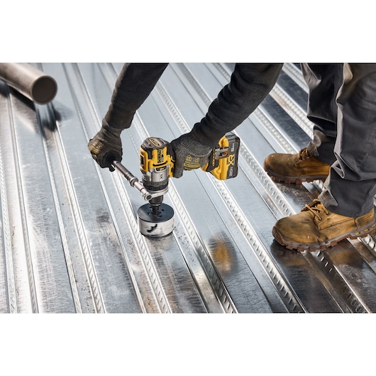 A person wearing work gloves and boots uses a DEWALT cordless drill to cut a hole in a shiny corrugated metal sheet on the floor.