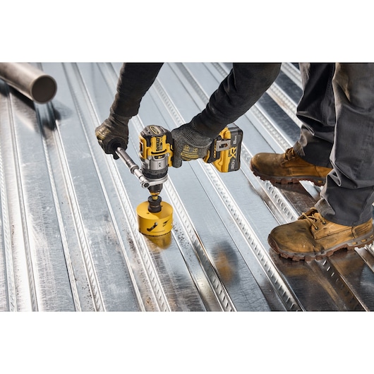 A person wearing work gloves and boots uses a DEWALT cordless drill with a hole saw attachment to drill into a corrugated metal surface.